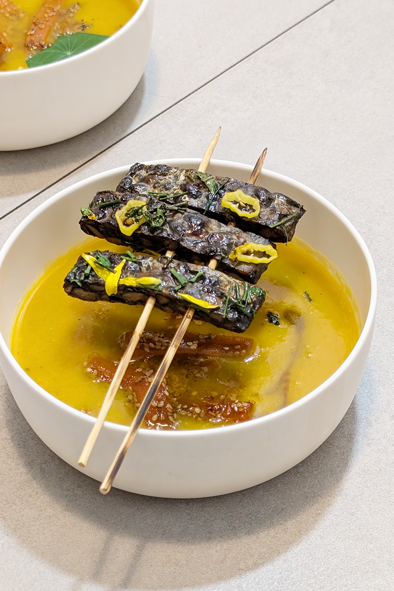 Vegane Möhrensuppe mit schwarzen Bohnen-Tempeh, serviert in einer Schüssel mit Kräutern.